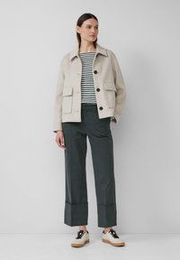 Beige wollen jas met twee voorzakken, donkere groene wijde broek en witte sneakers met zwarte accenten, gelaagd over een gestreept shirt.