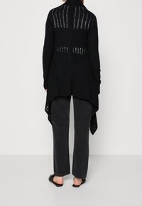 Svart stickad cardigan med öppen rygg, ribbad textur, lång längd och asymmetrisk fåll. Kombinerad med mörka byxor och platta sandaler.