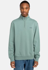 Mint green zip-neck sweatshirt made of soft fabric, featuring a ribbed hem and cuffs, with text logo printed on the left chest.