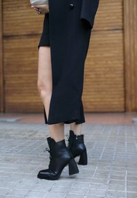 Person iført sorte læder snørebånds-ankelstøvler og en lang sort frakke med høj slids, der står på flisebelagt fortov foran trædøre.