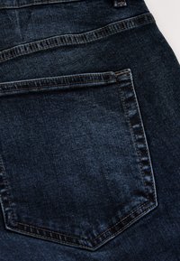 Jean en denim bleu foncé avec une poche arrière bien visible, des coutures discrètes et une finition propre avec de légères variations de texture.