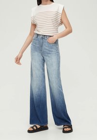 Žena ve bílo-beige pruhovaném topu bez rukávů, s vysokým pasem a širokými modrými ombre džíny a černými sandály, stojící s jednou rukou v kapse.