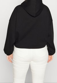 Czarna krótka bluza z kapturem, gładka w dotyku, z obniżonymi ramionami, zaprezentowana z białymi dopasowanymi spodniami. Prosty design, brak widocznych logo.
