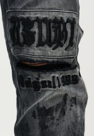 Γκρι distressed τζιν από denim με μαύρα κεντημένα γράμματα. Διαθέτει μεγάλες σκισίματα, αντίθετες υφές υφάσματος και ξεθωριασμένες περιοχές για μια φθαρμένη εμφάνιση.