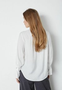 Hvid langærmet bluse med tekstureret stof, buet kant og knaplukning ved ærmerne. Modellens hår falder over skuldrene, iført grå bukser.