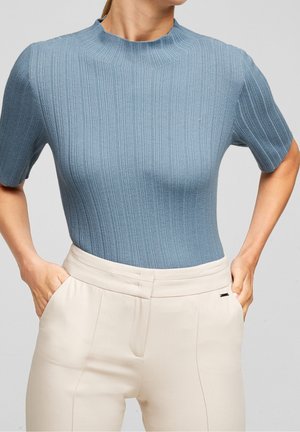 Kvinde iført en tætsiddende kortærmet ribbet blå sweater, der er stoppet ned i højtaljede cremede bukser med sidelommer, mens hænderne hviler i lommerne.