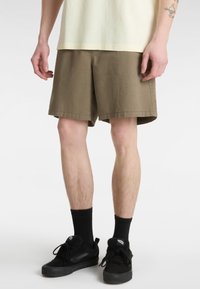 Bavlněné khaki šortky s volnějším střihem, bočními kapsami a lemem končícím nad kolenem, doplněné černými teniskami a ponožkami.