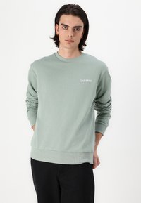 Lys grøn sweatshirt med ribstrik på manchetter og kant, med hvid tekst "Club1990" på venstre side. Modellen har sorte bukser på.
