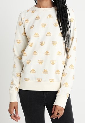 Frau trägt ein cremefarbenes Kappa-Sweatshirt mit wiederholten goldenen Logos, kombiniert mit schwarzen Jeans, posiert mit entspannt an den Seiten hängenden Armen.