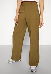 Kakifärgade byxor med vida ben och en slät textur, med flat front, sidfickor och medelhögt midjeanpassning, tillsammans med grå sportskor.