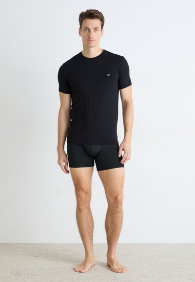 Tricou scurt negru cu mânecă scurtă și pantaloni scurți negri ajustați, confecționați dintr-un material neted. Tricoul are un mic emblema pe piept.