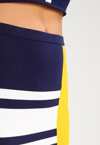 Námořnický modrožlutý pruhovaný crop top s bílými akcenty; přiléhavý design; hladká textura; kontrastní barvy vytvářejí geometrické vzory.