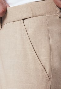 Beżowe spodnie materiałowe o teksturowanej tkaninie, z prostym przodem, kieszeniami bocznymi i zdobieniami wzdłuż talii oraz kieszeni.