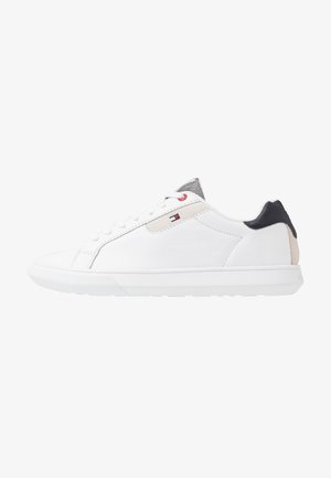 Baskets basses blanches avec lacets blancs, empiècement noir au talon, détail beige, et un petit logo de drapeau rouge, blanc et bleu sur le côté.