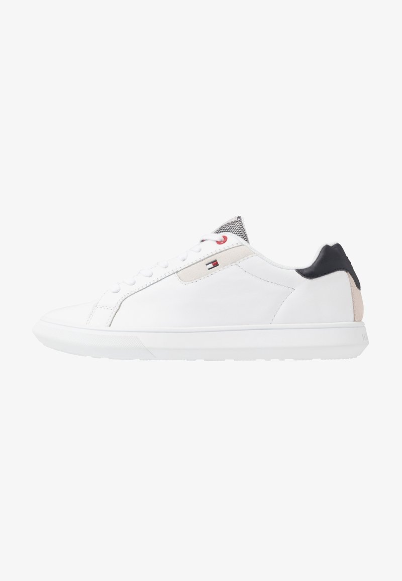 Baskets basses blanches avec lacets blancs, empiècement noir au talon, détail beige, et un petit logo de drapeau rouge, blanc et bleu sur le côté.