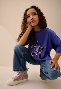 Camiseta de manga corta morada con diseño de estrella de lentejuelas. Jeans de pierna ancha azul desvaído. Zapatillas altas brillantes en colores pastel.