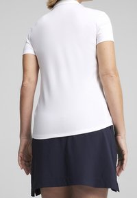 Kvinna står med ryggen mot kameran, iklädd en vit kortärmad polo och en marinblå kjol mot en enkel bakgrund.