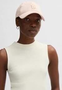 Rosa basebollkeps med broderad logotyp, tillverkad av bomull, med böjd skärm. Modellen bär en ärmlös ribbad topp i ljus krämfärg.