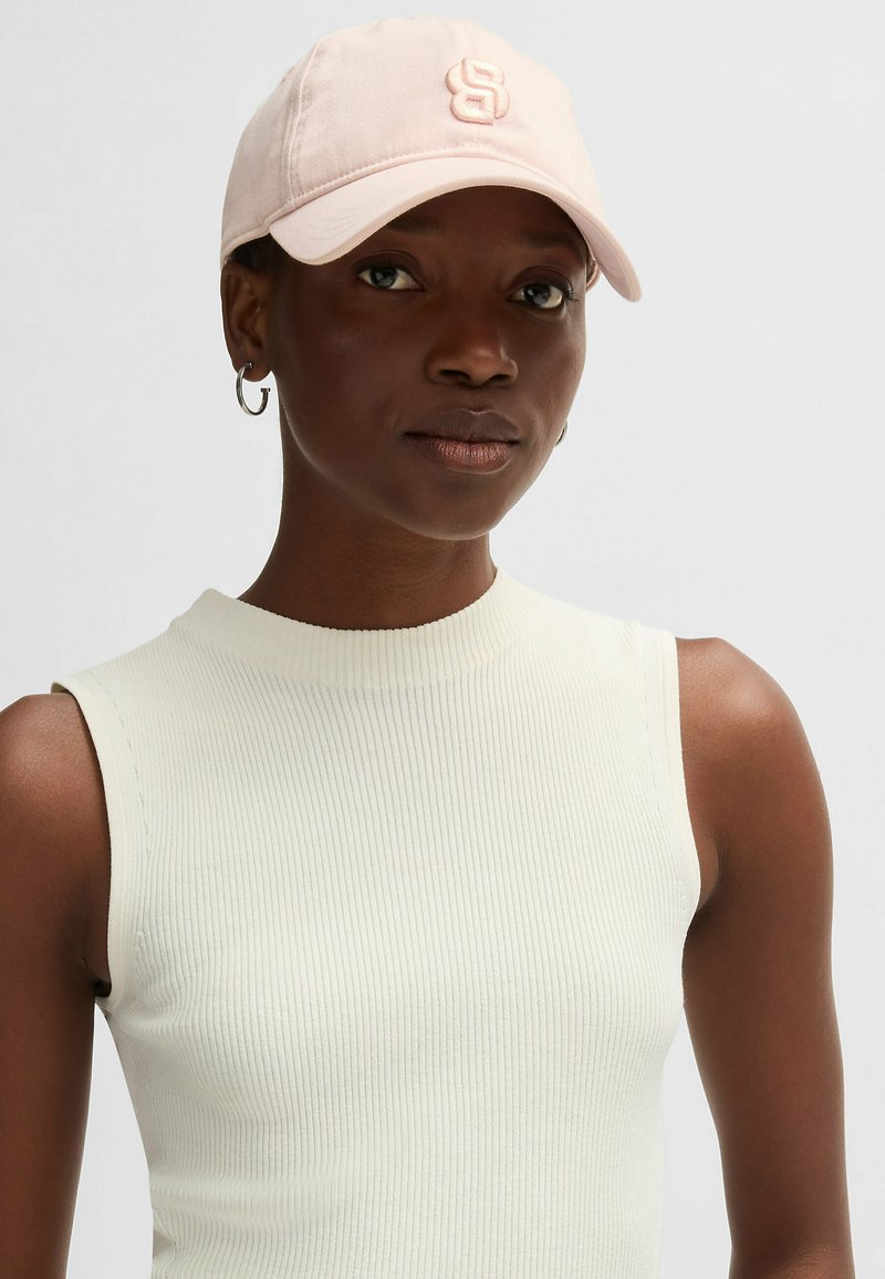 Rosa basebollkeps med broderad logotyp, tillverkad av bomull, med böjd skärm. Modellen bär en ärmlös ribbad topp i ljus krämfärg.