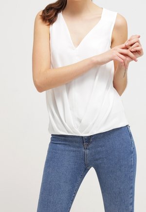 Blusa bianca senza maniche con scollo a V, caratterizzata da un dettaglio anteriore drappeggiato, abbinata a jeans di denim blu. Tessuto morbido e vestibilità comoda.
