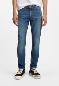 Μοντέρνο στενό τζιν παντελόνι από μπλε denim με ξεβαμμένο πλύσιμο, πέντε τσέπες και ραφές σε αντίθετο χρώμα. Συνδυασμένο με μαύρα αθλητικά παπούτσια με λευκές σόλες.