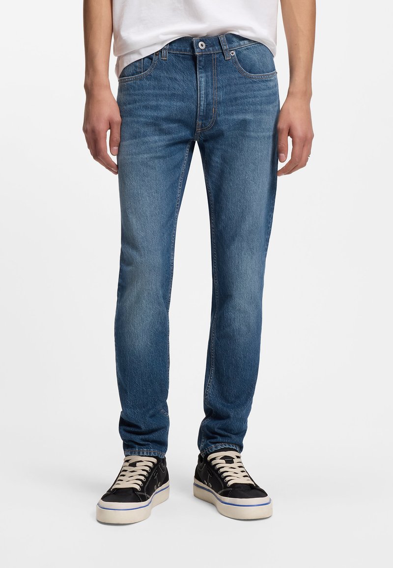 Μοντέρνο στενό τζιν παντελόνι από μπλε denim με ξεβαμμένο πλύσιμο, πέντε τσέπες και ραφές σε αντίθετο χρώμα. Συνδυασμένο με μαύρα αθλητικά παπούτσια με λευκές σόλες.