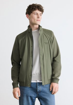 Ung mand iført en olivengrøn lynlåstrøje over en lysegrå sweater og blå jeans, kigger sidelæns mod en ensartet hvid baggrund.