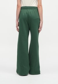 Άτομο που φοράει φαρδιά σκοτεινά πράσινα παντελόνια με ελαστική μέση, στέκεται με πλάτη σε ένα απλό λευκό φόντο.