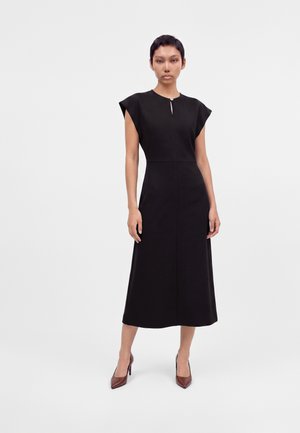 Femme aux cheveux courts portant une robe midi noire sans manches et des escarpins marron, debout devant un fond clair uni.