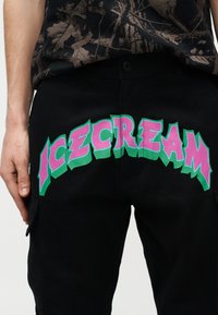 Personne portant un pantalon noir avec un grand texte stylisé « ICECREAM » en rose et vert dans le dos, assorti à une chemise sombre à motifs.