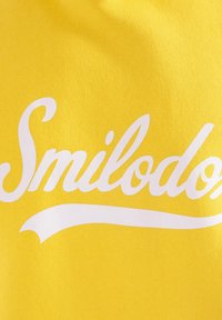Tessuto di cotone giallo con grande testo bianco che scrive "Smilodon" con una linea accentata curva sotto il testo.