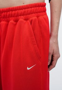 Punaiset collegehousut, joissa on teksturoitu kangas, joustava vyötärö, sivutaskut ja pieni valkoinen Nike-logo vasemmalla puolella.