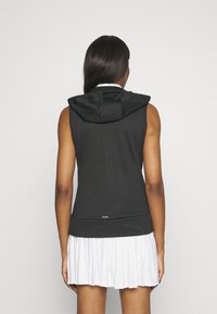 Sudadera sin mangas negra con un diseño ajustado, que incluye capucha, textura suave y un logo en el dobladillo. Combinada con una falda blanca con pliegues.