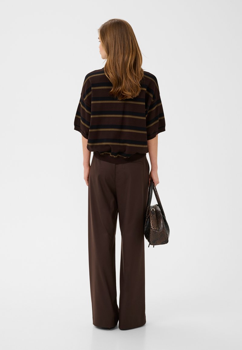 Femme aux longs cheveux bruns portant un haut ample rayé, un pantalon large marron, tenant un sac à main sombre texturé, debout, de dos, sur un fond blanc.