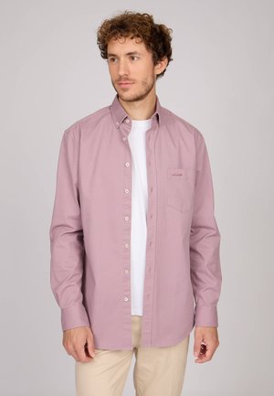 Chemise longue à manches longues de couleur violette claire avec col à boutons et poche plaquée, fabriquée en tissu lisse ; portée par-dessus un t-shirt blanc, associée à un pantalon beige.