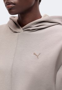 Gros plan d'une personne portant un sweat à capuche beige clair avec un petit logo Puma sur la poitrine gauche sur un fond uni.
