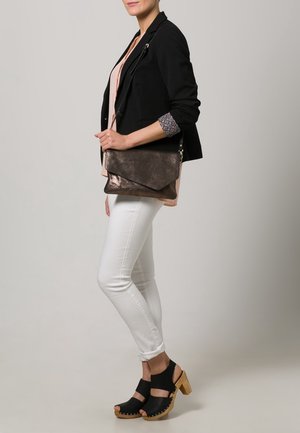 Zwarte blazer met een ritssluiting, lichtroze shirt, witte skinny jeans en zwarte klompen met een houten hak, terwijl ze een metalen donkerbruine tas draagt.