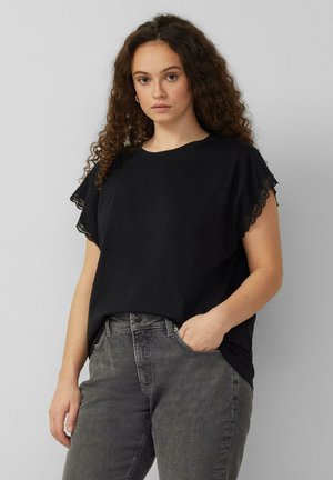 Junge Frau mit lockigem braunem Haar, die ein schwarzes, mit Spitze besetztes kurzärmeliges Oberteil und ausgewaschene graue Jeans trägt, vor einem hellgrauen Hintergrund stehend.