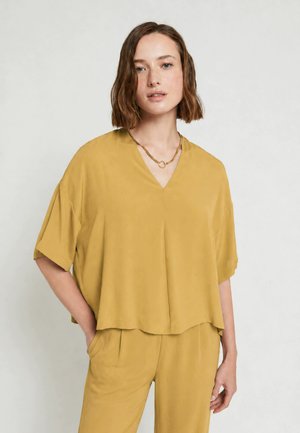 Donna con capelli corti e mossi che indossa una maglia larga color senape a scollo a V e pantaloni abbinati, in piedi con una mano in tasca.