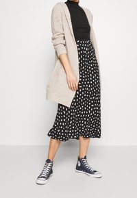 Długa czarna spódnica z białym i brzoskwiniowym abstrakcyjnym wzorem, zestawiona z czarnym golfem i jasnobeżowym, dzianym kardiganem. Niebieskie trampki na wysokiej podeszwie.