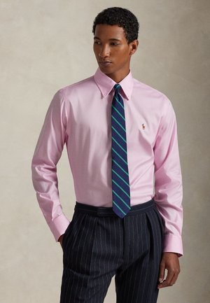 Άνδρας που φοράει ροζ πουκάμισο με μικρό λογότυπο, γραβάτα με μπλε και πράσινες ρίγες και σκοτεινό ριγέ παντελόνι σε ουδέτερο φόντο.