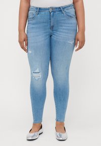 Light blue skinny jeans with distressed detailing on the thighs, five-pocket design, and a button-front closure. Worn with silver flats.