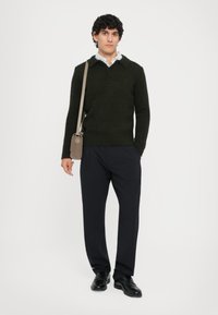 Dunkelgrüner, gestrickter Pullover mit einem Kragen, kombiniert mit lockeren, schwarzen Hosen und schwarzen Schuhen; kleine beigefarbene Umhängetasche.