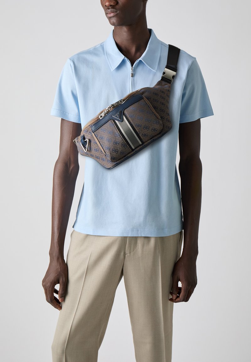 Bolso de hombro marrón con un diseño estampado y un acento azul, que cuenta con un cierre de cremallera, herrajes metálicos y una correa ajustable negra. Lleva puesto sobre una camisa azul claro.