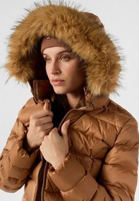Bruine, gewatteerde jas met een capuchon gevoerd met bont, een ritssluiting en een gestructureerde stof met een doorgestikt ontwerp. Model houdt de kraag vast.