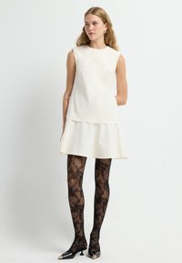 Crème mouwloze jurk met gelaagde zoom, gecombineerd met zwarte sheer kant panty en glanzende zwarte hakken met een spitse neus en gouden accenten.