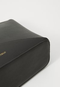 Coin d'un sac en cuir noir rectangulaire avec une couture subtile et un texte de marque embossé en or partiellement visible sur un fond clair uni.