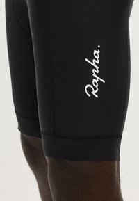Gros plan sur un short de cyclisme noir porté par une personne, avec le texte blanc « Rapha. » imprimé sur la cuisse.