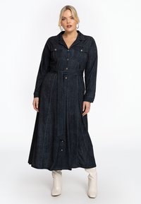Robe chemise en denim bleu marine avec manches longues, fermeture à boutons, deux poches poitrine, taille ceinturée et jupe fluide, associée à des bottes blanches.