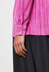 Blusa rosa plissettata con maniche lunghe, caratterizzata da un polso con bottoni. Il materiale ha una texture lucida e il design presenta strisce verticali.